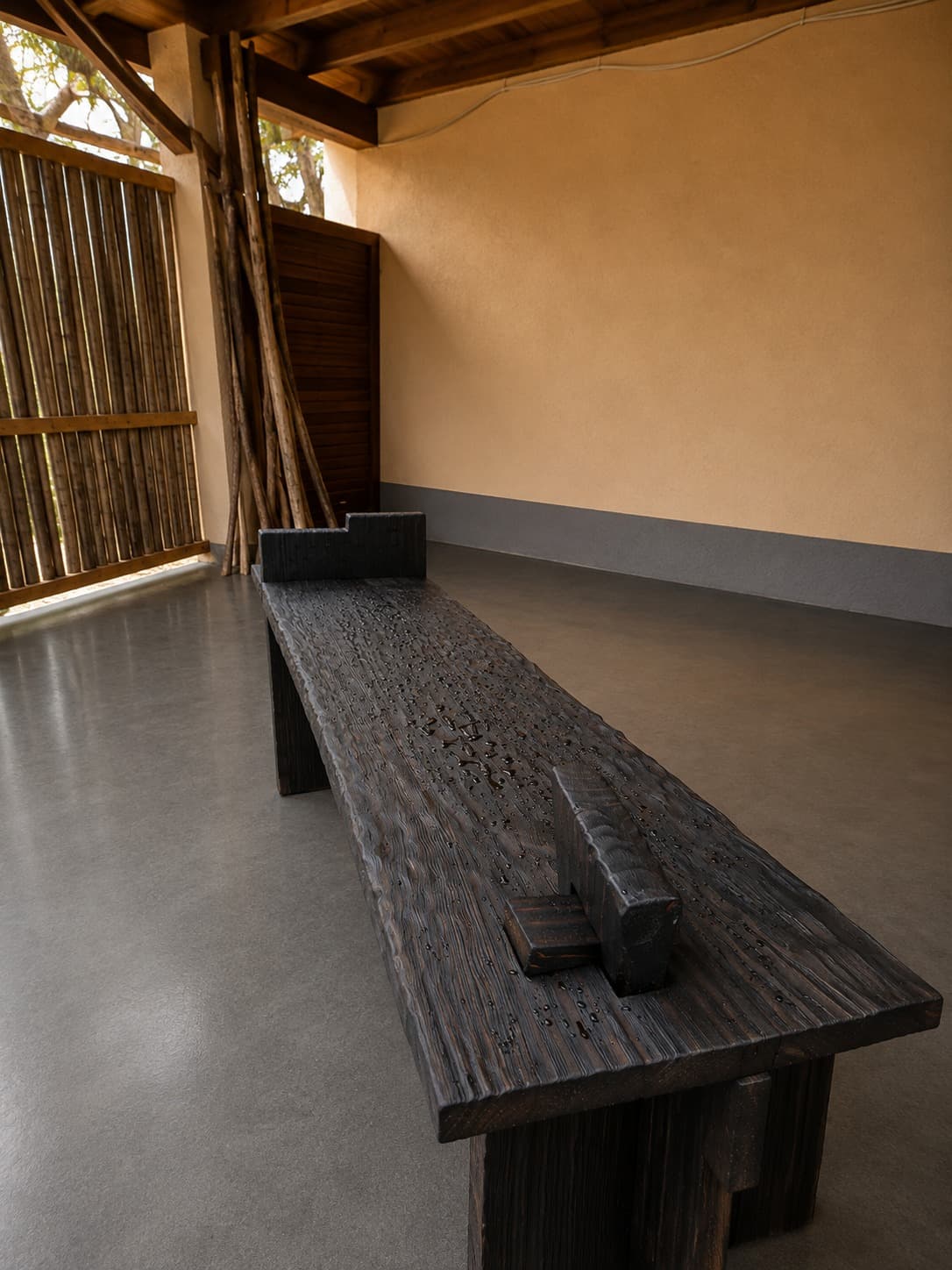 Mesa de arrime en madera quemada, de tabla tallada y patas encastradas, en una galería de muro ocre y piso de cemento.